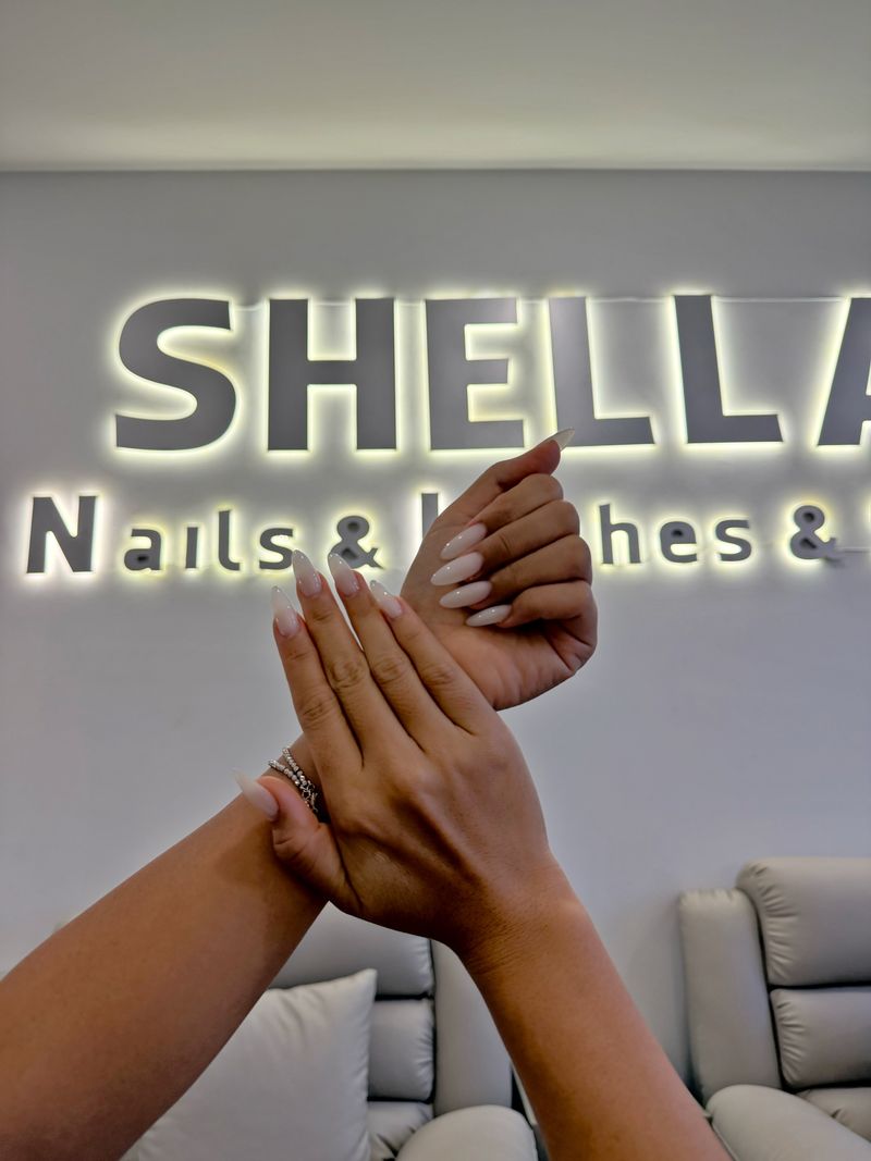 Long milky-white almond nails, shown in front of the Shella Nails & Lashes sign in Da Nang
